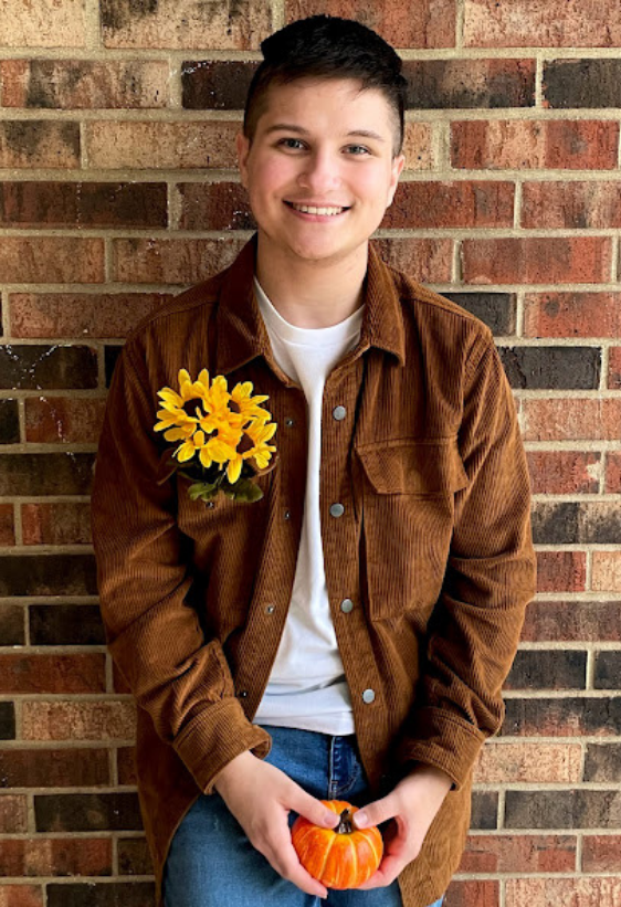 Picture of a smiling person with brown hair wearing a brown shirt standing in front of a brick wall
